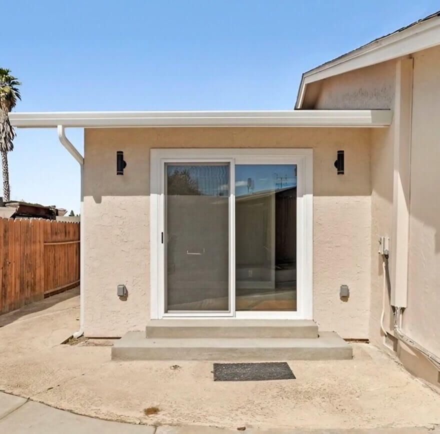 A clean, modern back entrance with sliding glass doors, flanked by wall sconces, leading to a backyard with a wooden fence.