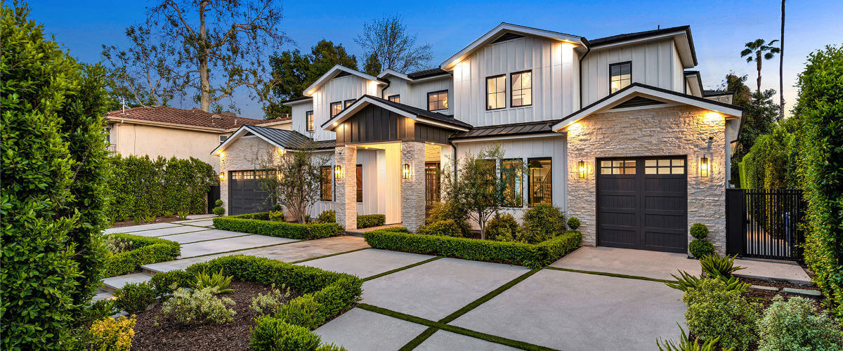 Modern two-story house with a stylish façade, manicured lawn, and landscaped driveway in twilight. Lush greenery borders the property.