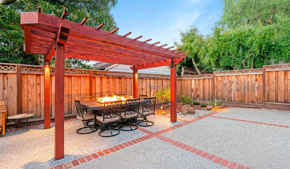 A cozy outdoor dining area under a wooden pergola, featuring a fire pit surrounded by black swivel chairs and a landscaped yard.