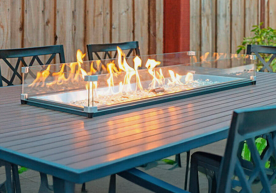 A glass-enclosed fire pit with flickering flames sits on a modern outdoor dining table surrounded by chairs.