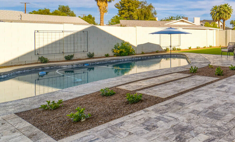 A serene backyard featuring a pool, patio with stone tiles, and a shaded area with an umbrella, surrounded by neatly arranged plants.