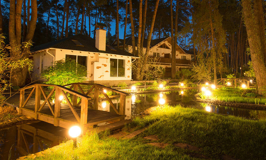 A serene nighttime view of a cozy house beside a pond, illuminated by soft lights and surrounded by tall trees.