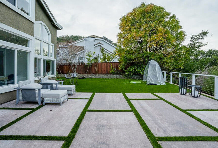A spacious patio features modern furniture, vibrant green grass, and a backdrop of a large tree and neighboring houses under a cloudy sky.