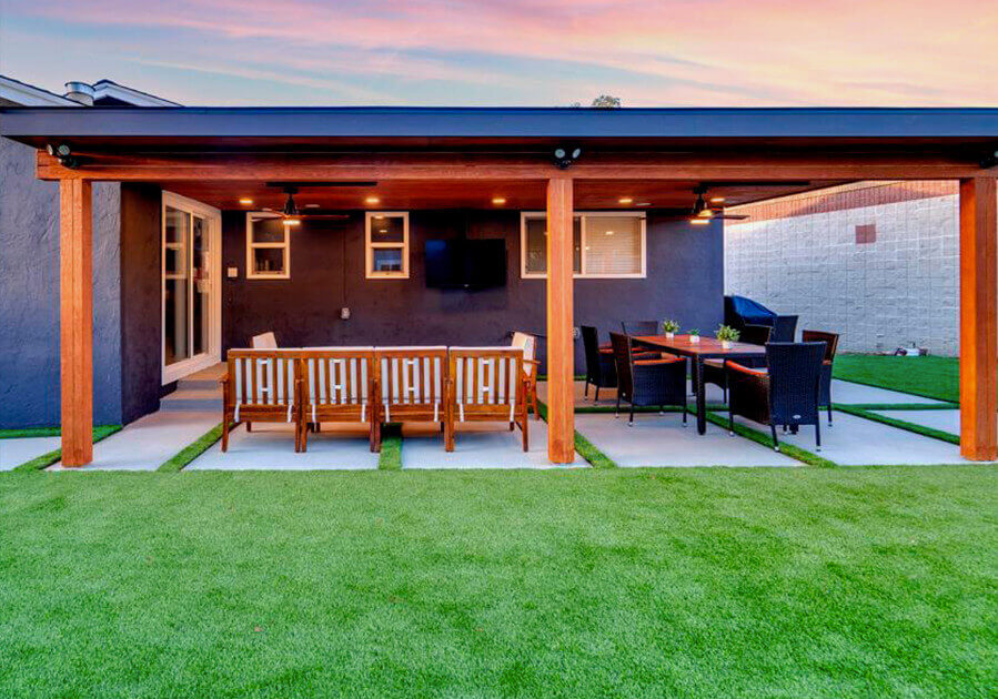 A stylish outdoor patio with wooden furniture, a dining table, and lush green grass under a colorful sunset sky.