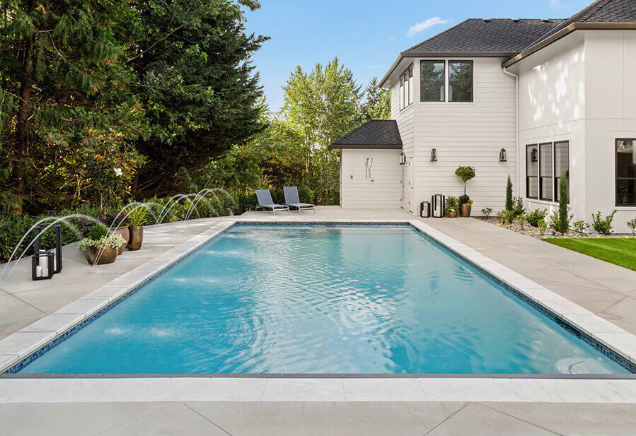 Backyard view featuring a sparkling pool with fountains, surrounded by greenery and a modern home. Two lounge chairs are nearby.