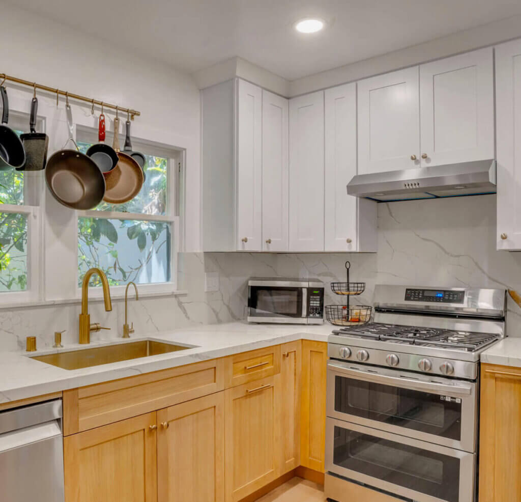 Modern kitchen featuring light wood cabinets, stainless steel appliances, a gold faucet, and hanging pots above a white marble countertop.