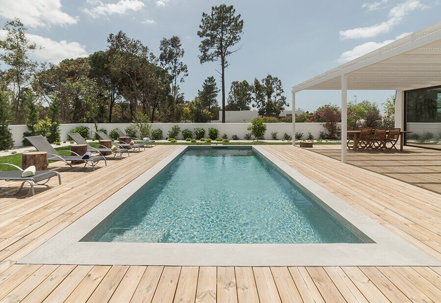 Modern outdoor space featuring a rectangular swimming pool, wooden deck, lounge chairs, and lush greenery under a clear sky.