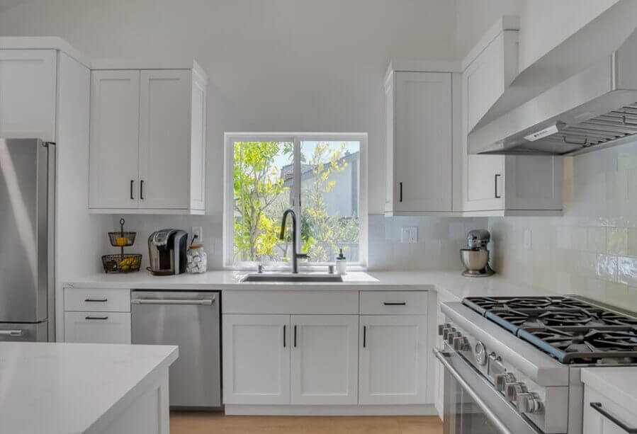 Modern white kitchen with stainless steel appliances, large window, and gas stove, featuring stylish cabinetry and natural light.
