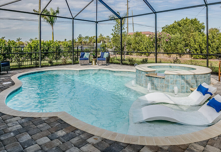 Pool area with a curved shape, spa, and lounge chairs under a screened enclosure, surrounded by lush greenery and blue skies.
