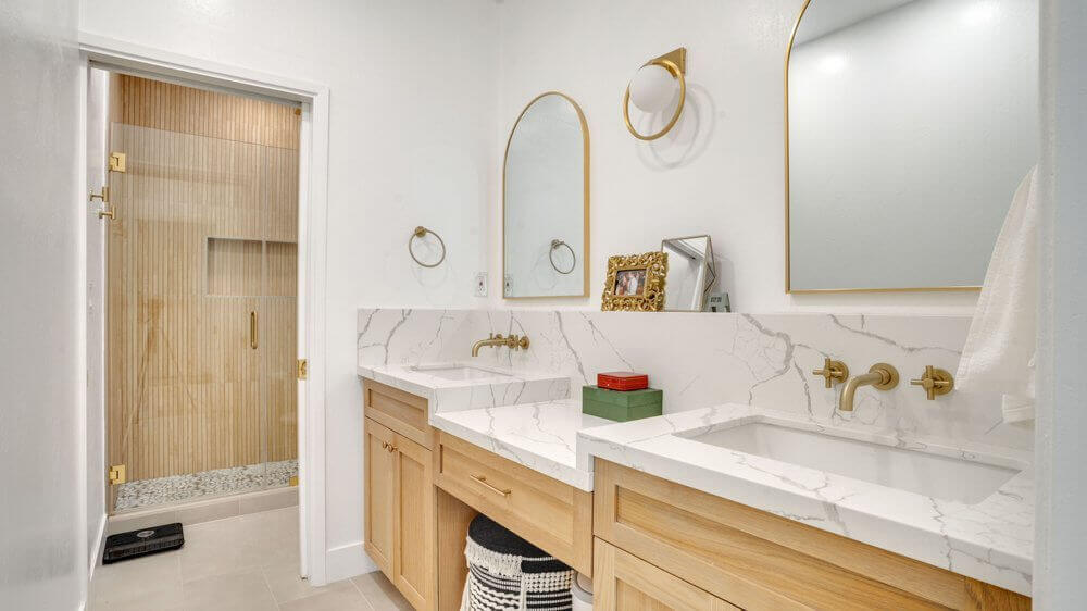 White marble bathroom with gold trim mirrors and light wood-colored drawers. A walk-in shower with bamboo walls.