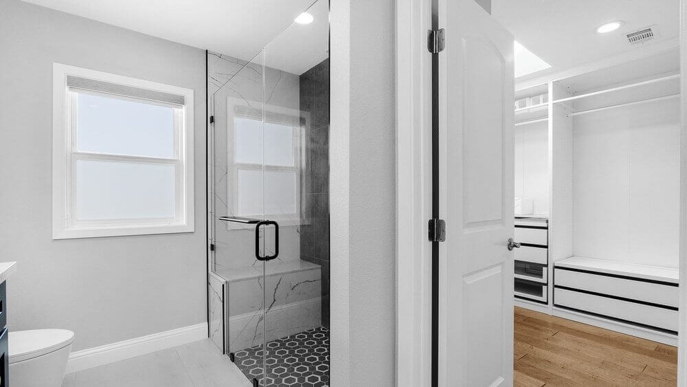 A white bathroom with a walk-in shower with a hexagonal pattern on the floor. There is a window in the bathroom, and the bathroom leads to a large walk-in closet with the same black and white color scheme.