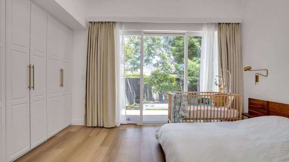 A bedroom with two sets of double door closets. There is a baby crib next to a white bed, and a glass sliding door leading to the backyard.